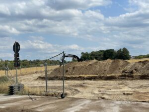 Blick auf Erdhaufen hinter eine Fläche von der die Erde abgetragen wurde. Llinks ein Tor aus bazäunen und im Hintergrund ein Bagger.
