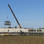 2023-10-11 Neubau 00001 Ein Autokran hebt ein Wandsegment über einem zweistöckigen Rohbau.