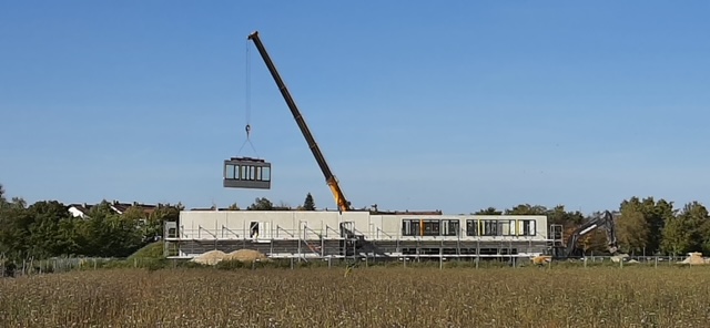 Ein Autokran hebt ein Wandsegment über einem zweistöckigen Rohbau.