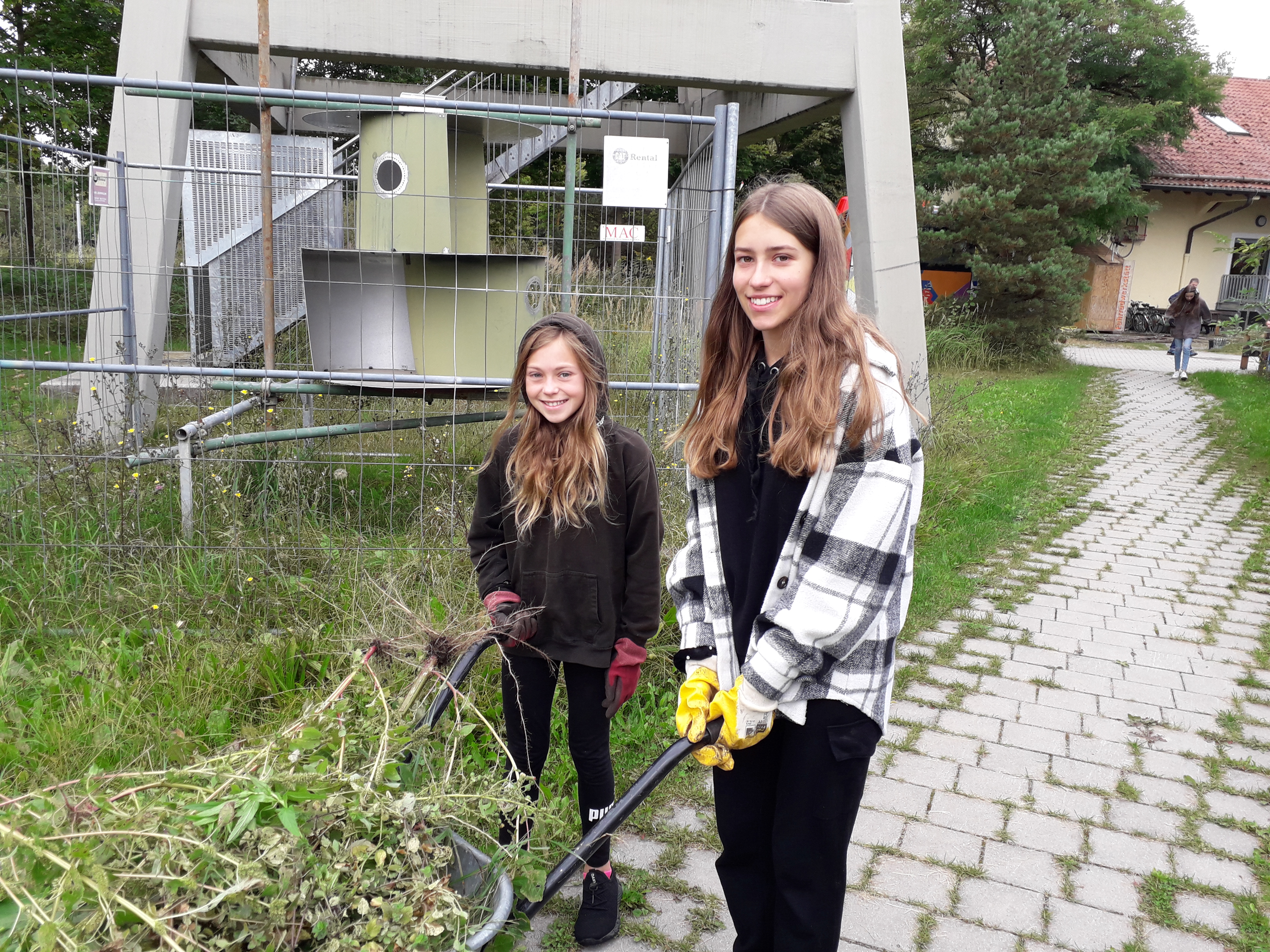 Zwei lächelnde Jugendliche mit einer mit Heckenschnitt gefüllten Schubkarre.